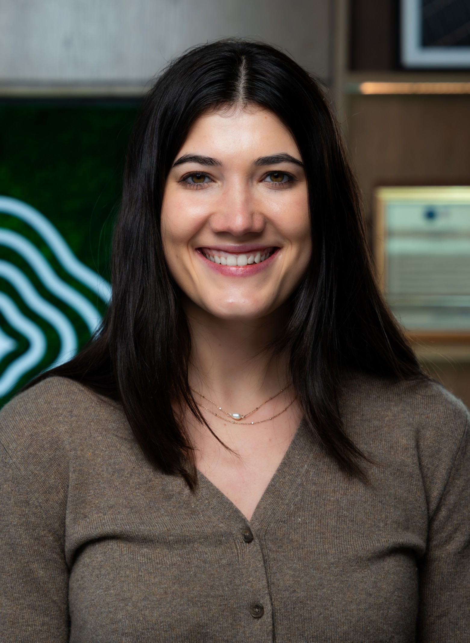 image_Rachel A woman with long dark hair, wearing a brown sweater and a necklace, smiles at the camera in an indoor setting with blurred wall decor in the background.