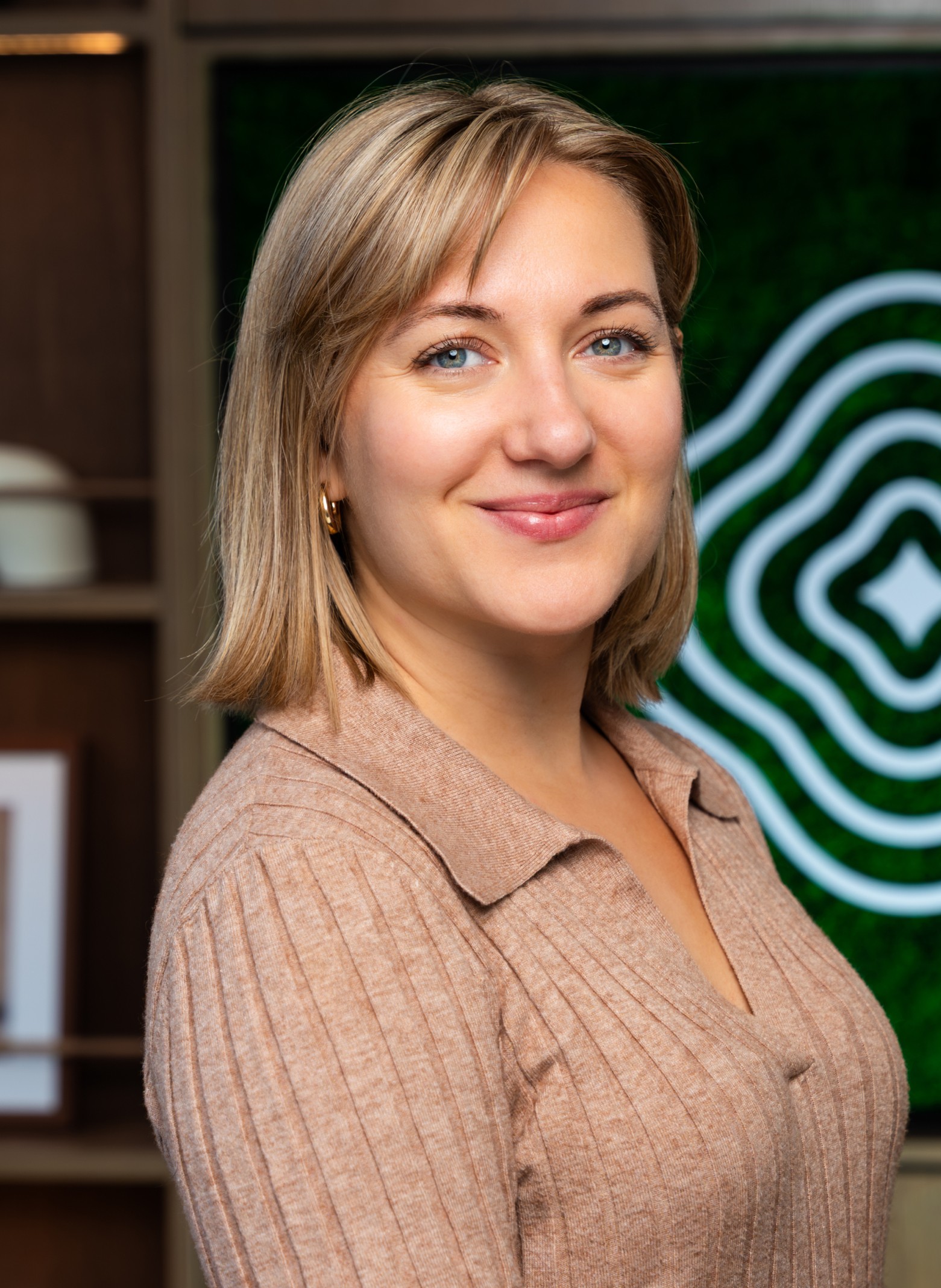 image_Sara A woman with blonde hair and blue eyes, wearing a tan ribbed shirt, smiles at the camera. She stands indoors with shelves and a green patterned wall in the background.
