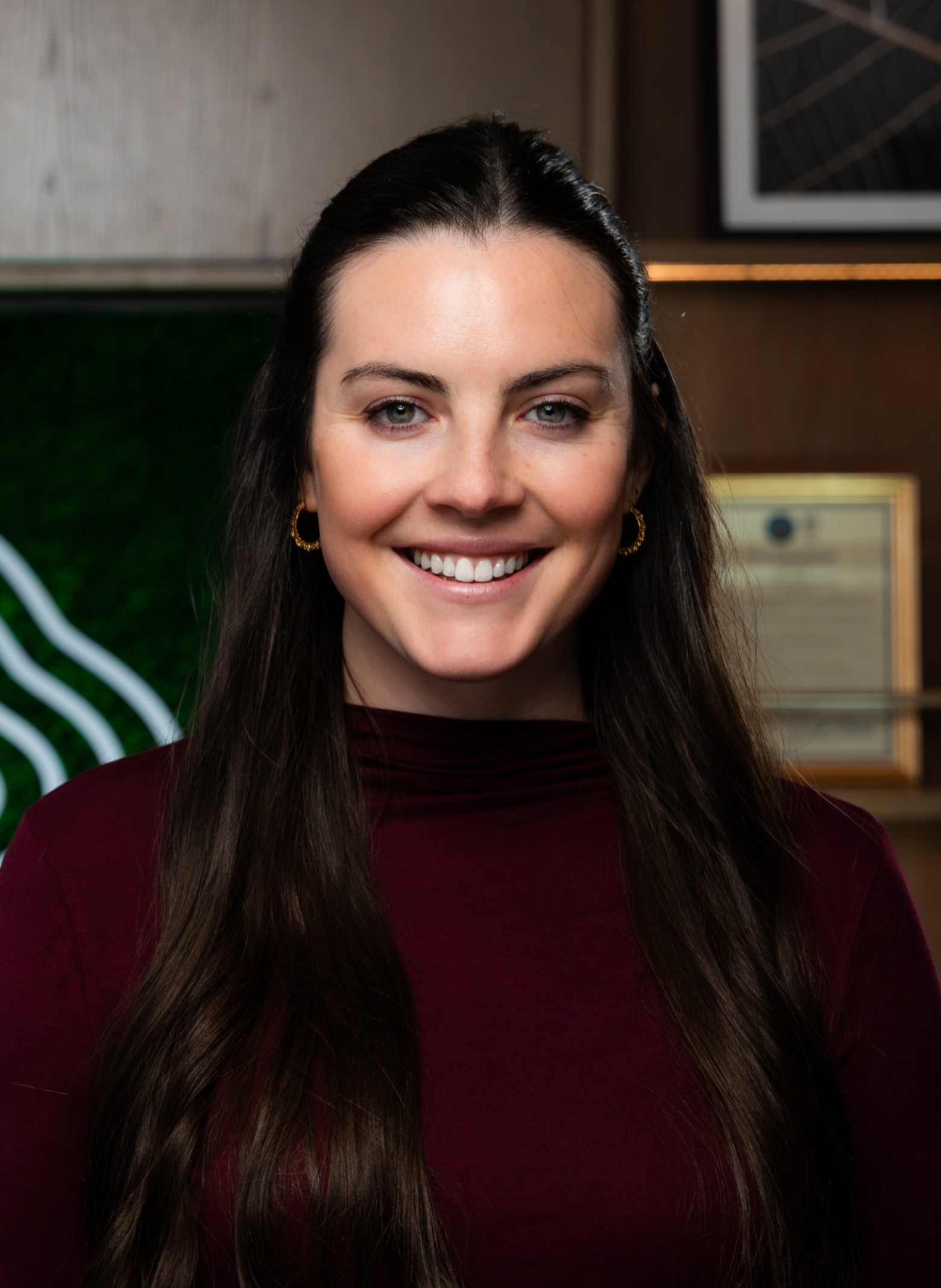 image_kaylee Kaylee LeCavalier wearing a burgundy top, smiles at the camera in an indoor setting with certificates and artwork in the background.