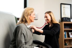A female healthcare professional smiles while using a stethoscope to listen to the chest of an older woman in a medical office, demonstrating the personalized care often provided by a concierge doctor. What is a Concierge Doctor?.