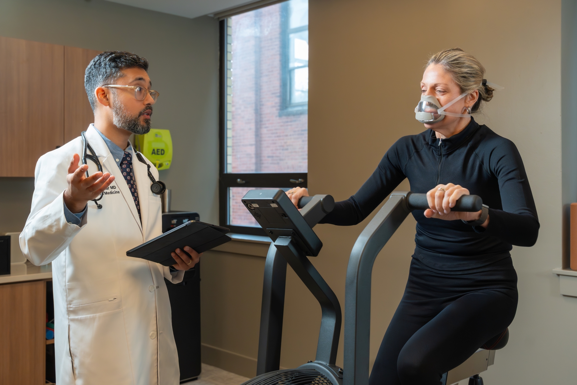 Dr. Luthar in a white coat speaks to a woman wearing a breathing mask while she exercises on a stationary bike in a medical office. The doctor holds a tablet, monitoring her performance.