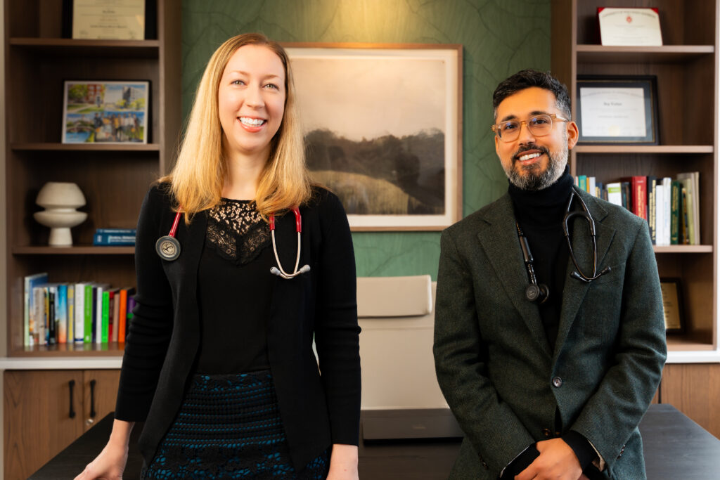 Two doctors, a woman and a man, stand smiling in an office with bookshelves, framed certificates, and a landscape photo on the wall behind them. Both wear stethoscopes around their necks.
