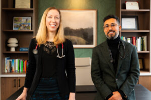Two doctors, a woman and a man, stand smiling in an office with bookshelves, framed certificates, and a landscape photo on the wall behind them. Both wear stethoscopes around their necks.