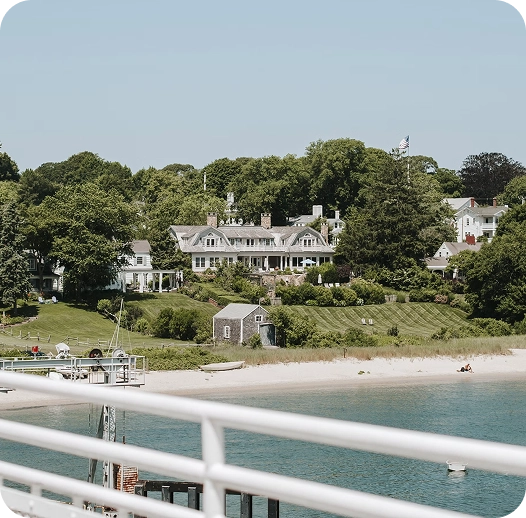 hero-primary-large-resort Large waterfront house surrounded by lush trees and greenery, with a sandy beach and calm blue water in the foreground. A white railing runs across the bottom of the image.