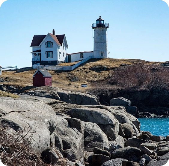 hero-primary-lighthouse-2 A white lighthouse and adjacent house sit on a rocky, grassy hill by the ocean, with a small red shed nearby and blue sky overhead.