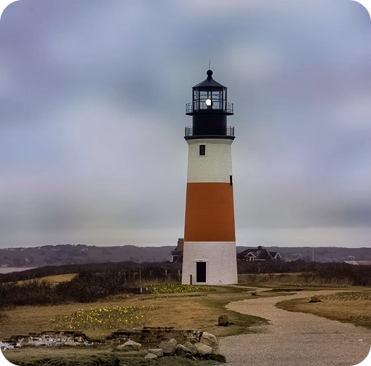 hero-primary-lighthouse A tall lighthouse with a white base, orange center, and black top stands on a grassy landscape under a cloudy sky, with a winding path leading up to it.