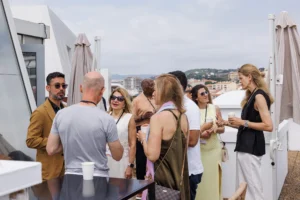 A group of people stand and chat on a rooftop terrace on a sunny day, embodying the idea that 80% of health and wellness comes from social connection and spending time outdoors. Some hold cups, and the cityscape is visible in the background. The group appears to be enjoying a casual outdoor gathering.