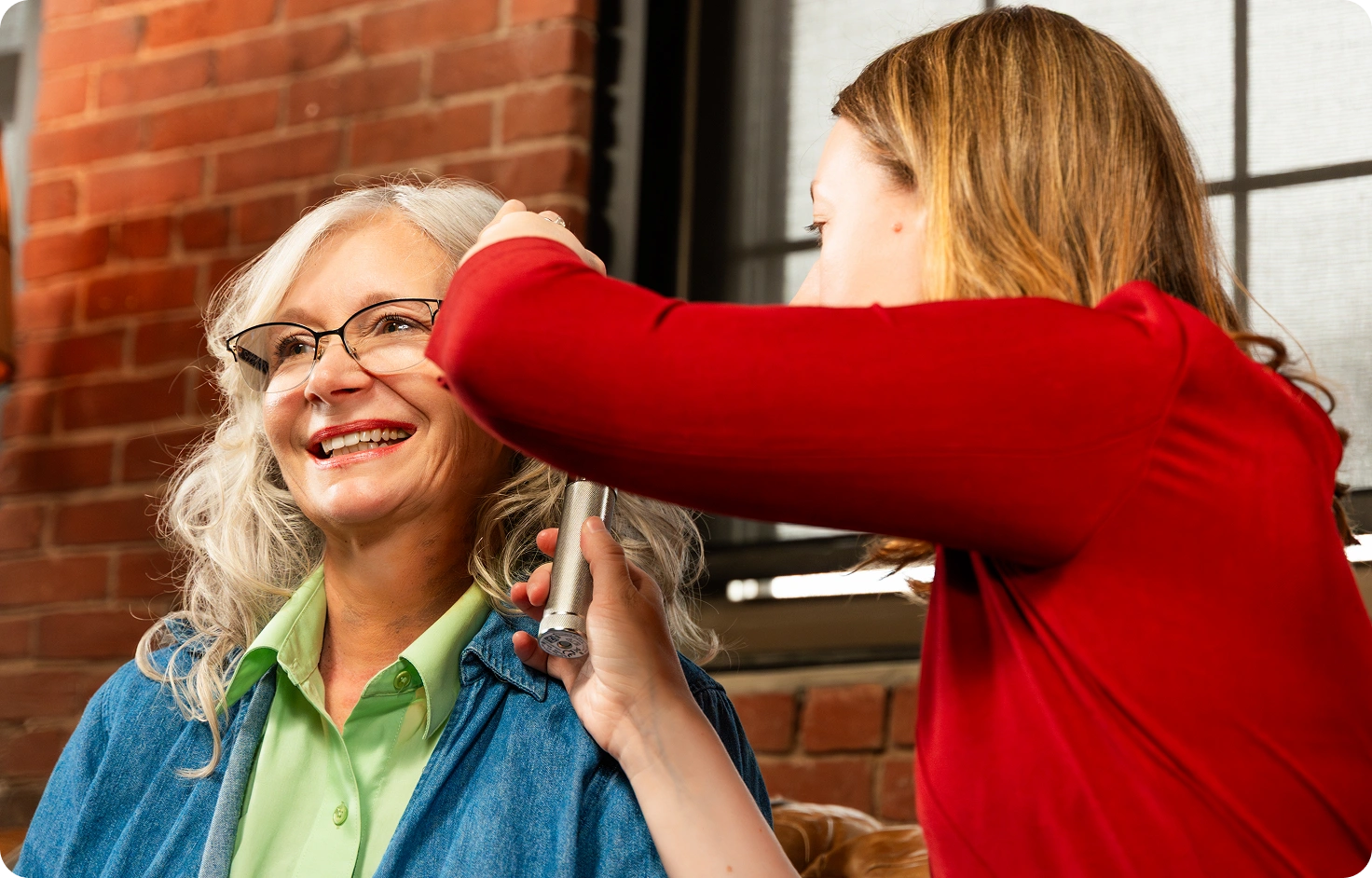 who-right-sticky-woman-getting-ear-check A smiling older woman with gray hair and glasses gets her hair styled by another woman in a red top, against a brick wall background.